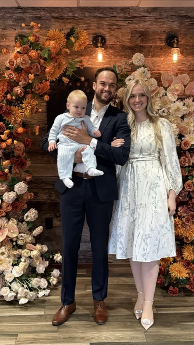 A young family with their baby in front of a wall of autumn flowers